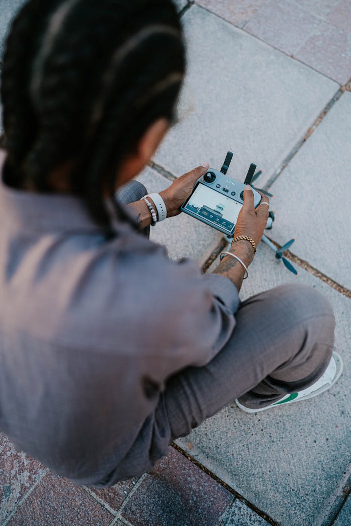 A young individual controlling a drone with a remote, seated on paved ground outdoors.